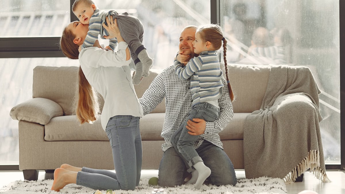 Cheerful mother kissing little boy while father hugging adorable daughter when family sitting on floor in rainy day against window